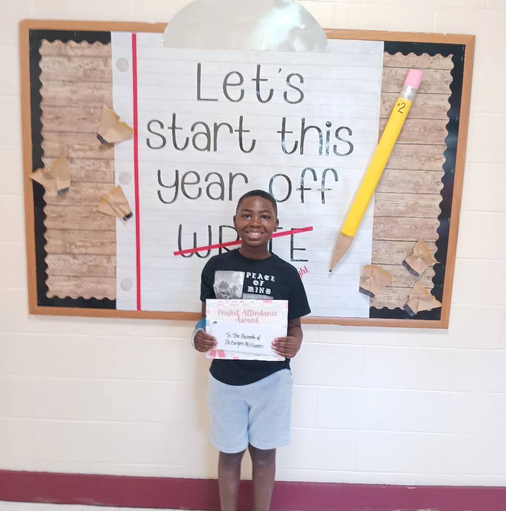 A student holds a certificate and stands in front of a sign that says "Let's start this school year off write", with write crossed out and replaced with "right."