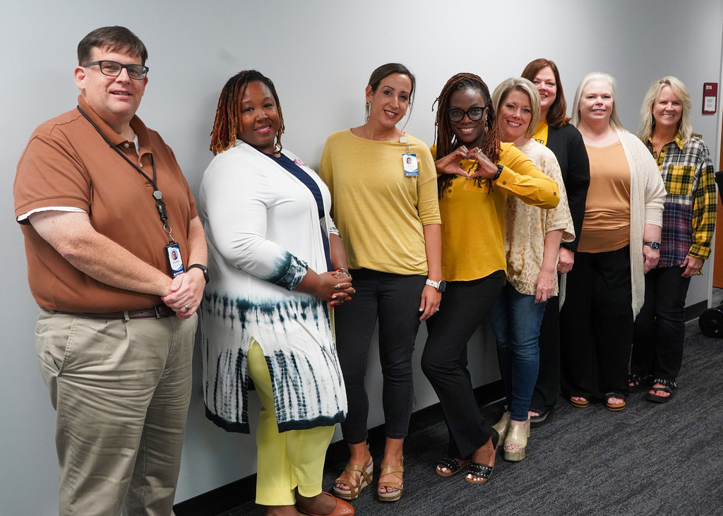 7 women and 1 man stand together in a hallway and pose for a photo. They’re all wearing gold for Childhood Cancer Awareness.