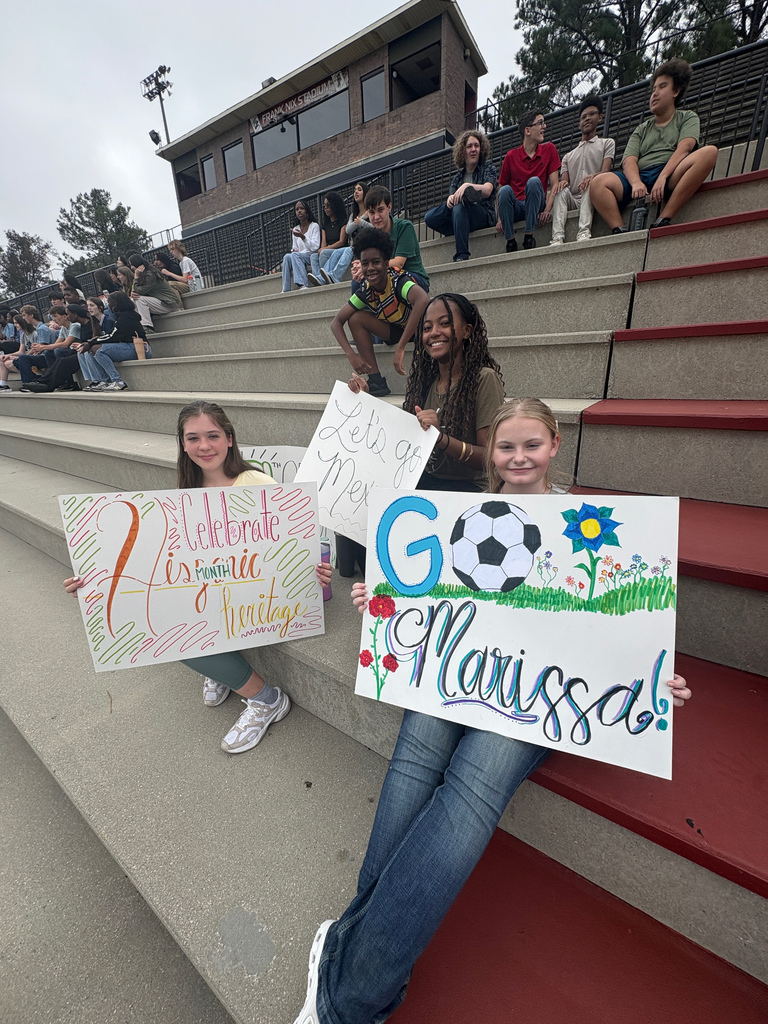 Students sit in stands, watching a soccer game and holding signs. One sign says "Celebrate Hispanic Heritage Month." Another says "Let's go Mexico." Another sign says "Go Marissa!"