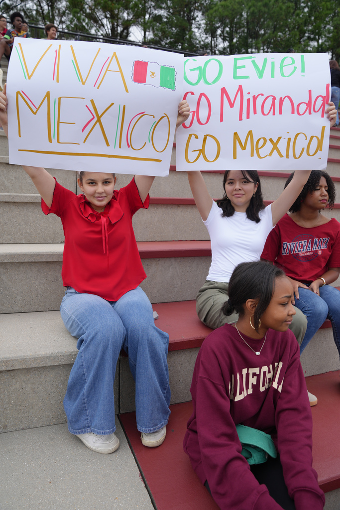 Girls sitting in stands hold up signs. One says "Viva Mexico." The other says, "Go Evie! Go Miranda! Go Mexico!"