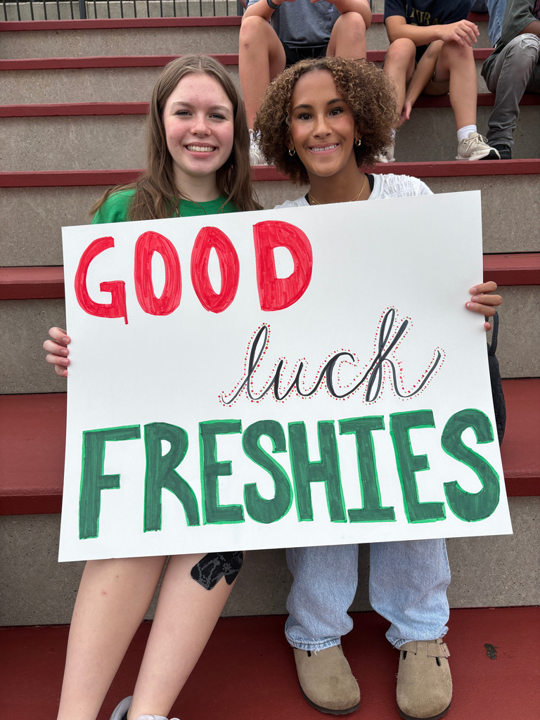 Two girls sit in stands, holding a sign that says "Good Luck Freshies"