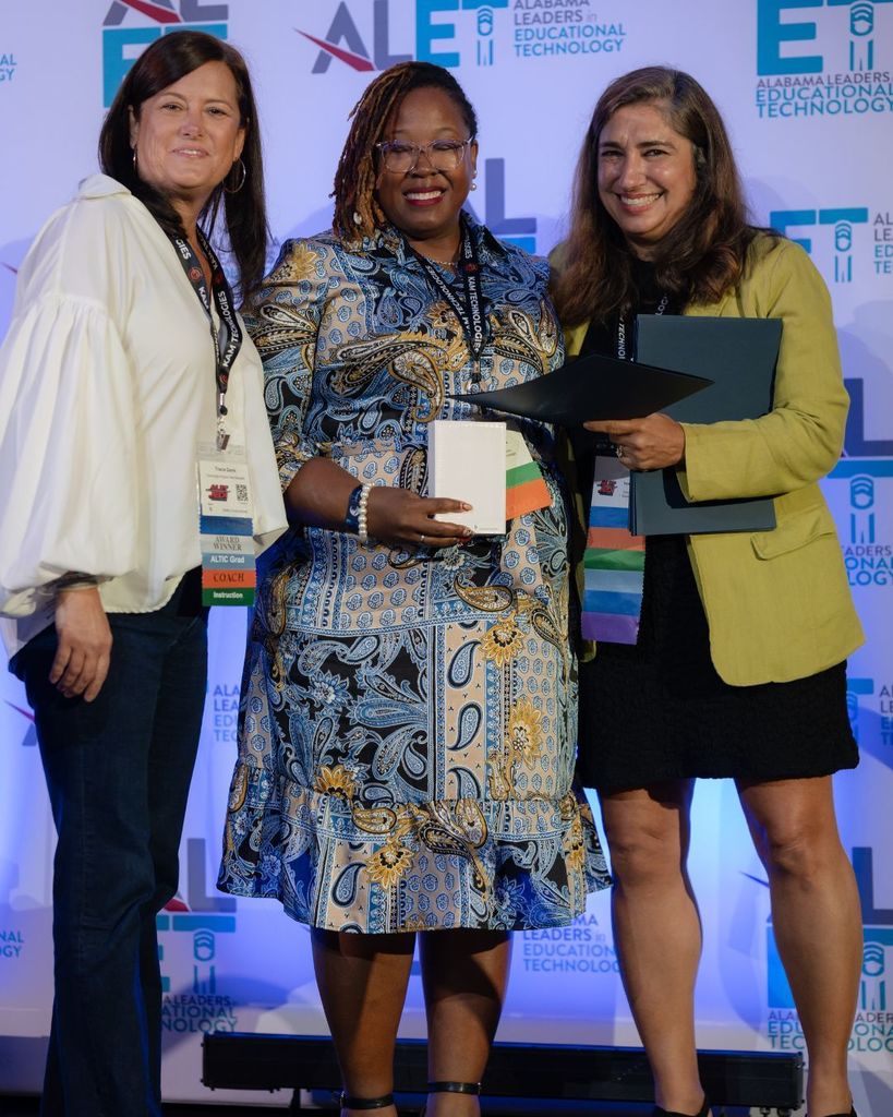 Three women stand together on a stage. Two of the women hold awards.
