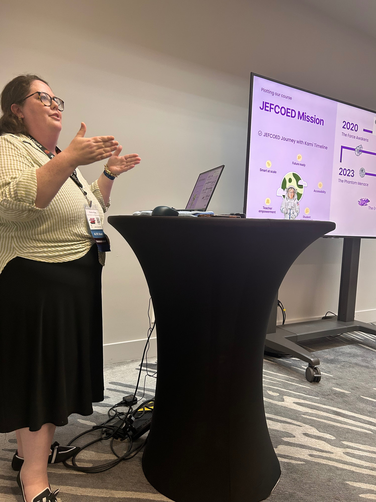 A woman stands in the front of a room, giving a presentation.
