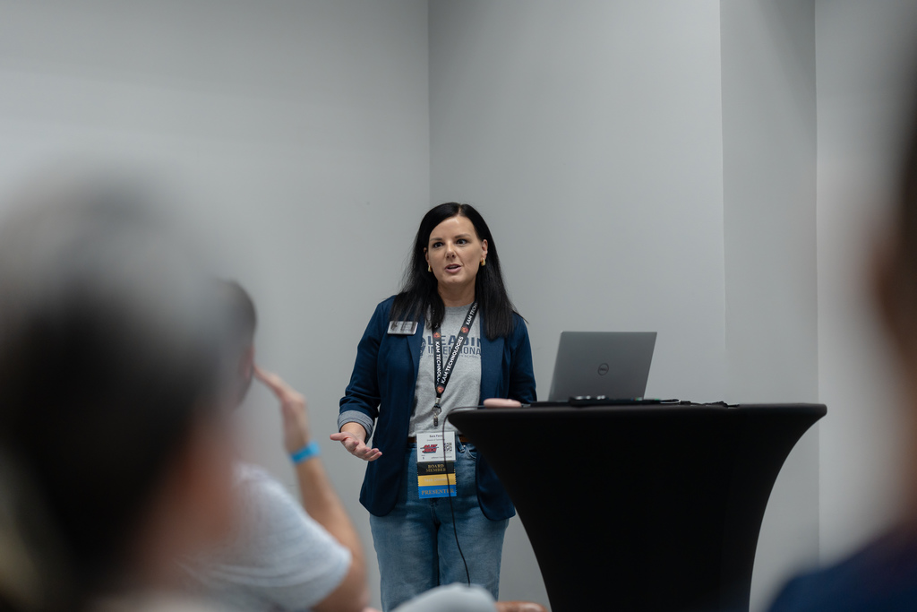 A woman stands in the front of a room, giving a presentation.