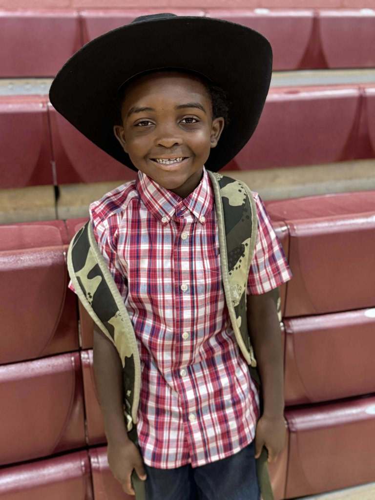 A little boy wearing a flannel shirt and cowboy hat smiles while standing in a school gym.