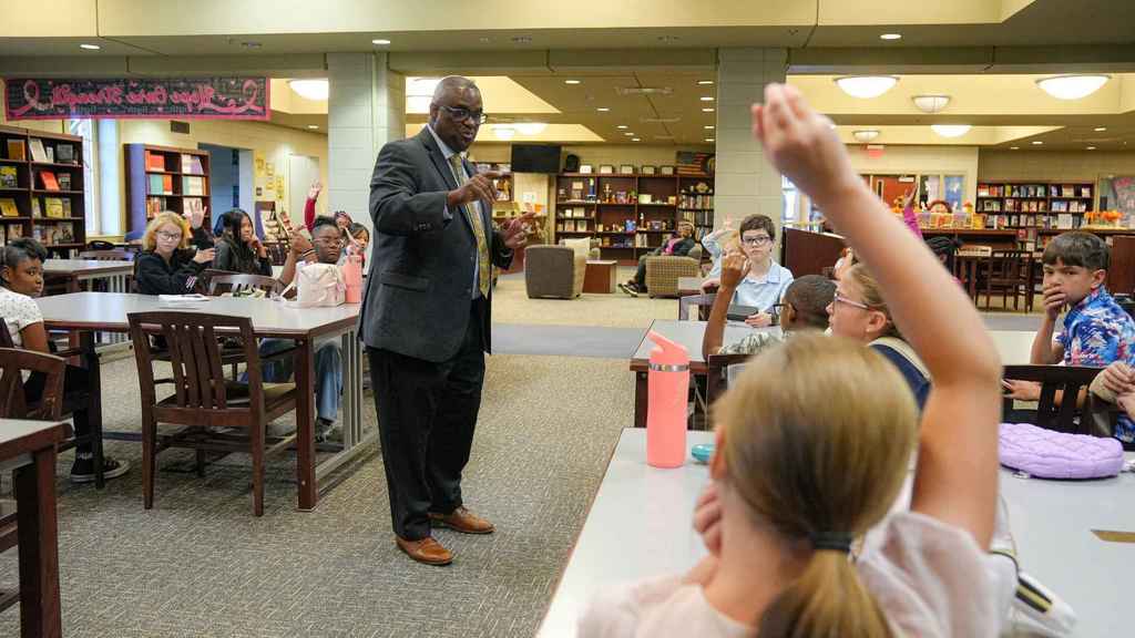 JEFCOED Superintendent Dr. Gonsoulin stands in a library and talks to children. The students sit at tables in the library. Some have their hands raised.