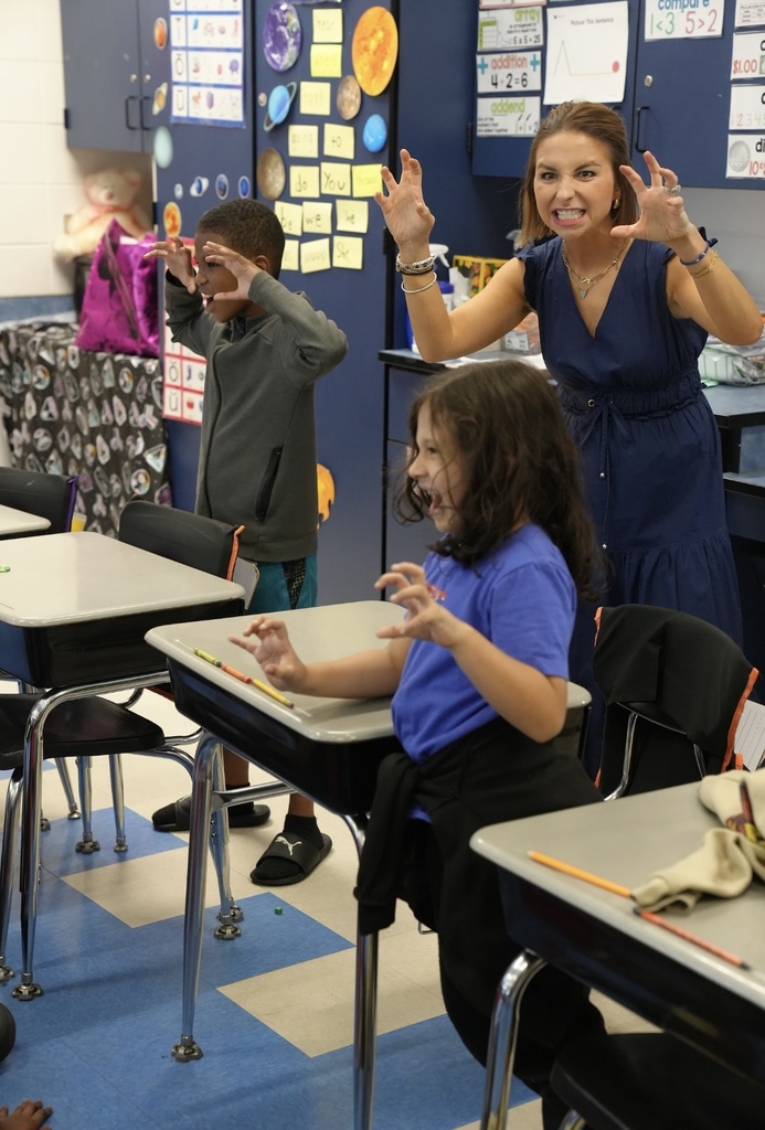 Students and an adult are in a classroom, practicing theatre techniques. They all hold their hands up and make scary faces.