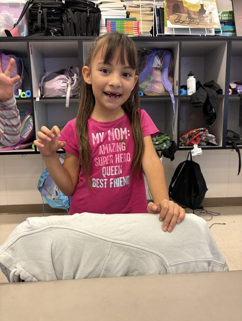 A student stands in a classroom, holding one of her hands up and smiling.