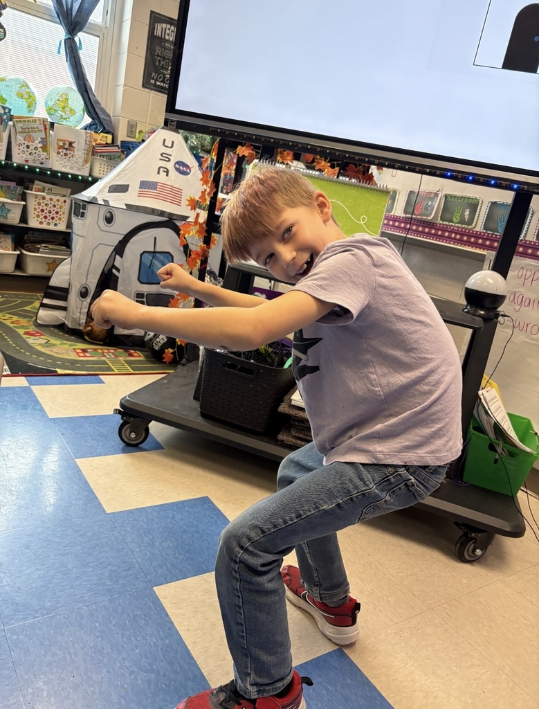 A student stands in a classroom, acting out an assignment to create a tableau of a bicycle. The student is in a squated position, holding his arms out.