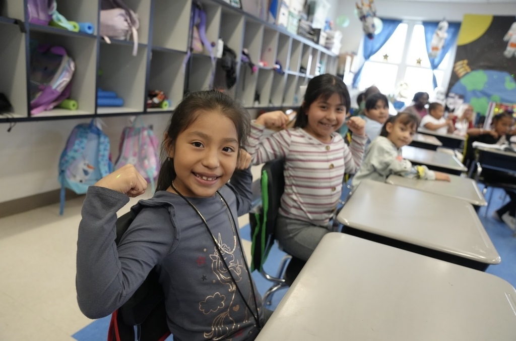 Students sit in a classroom, holding their arms up in a flexed position and smiling.