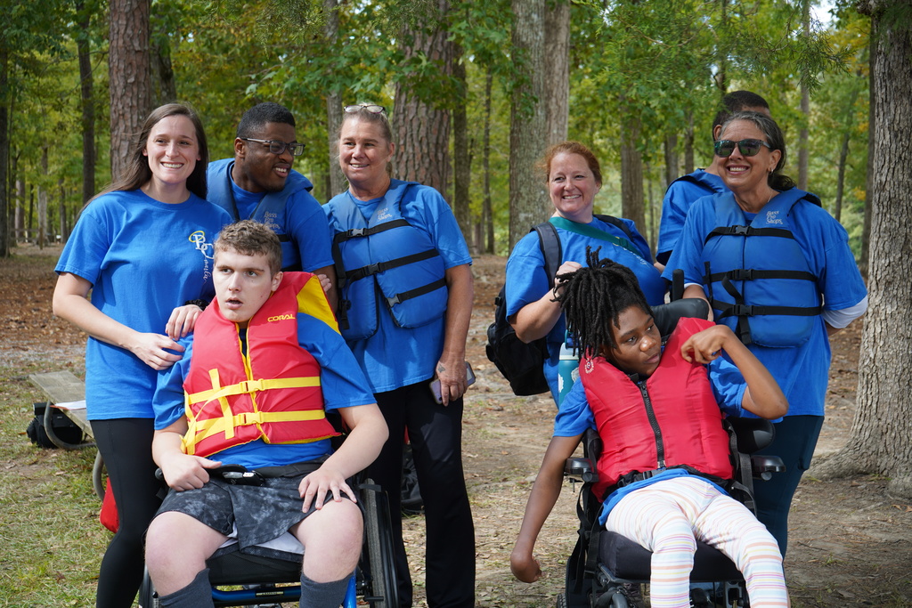 Students and teachers stand and sit together, smiling for a photo in a wooded area.