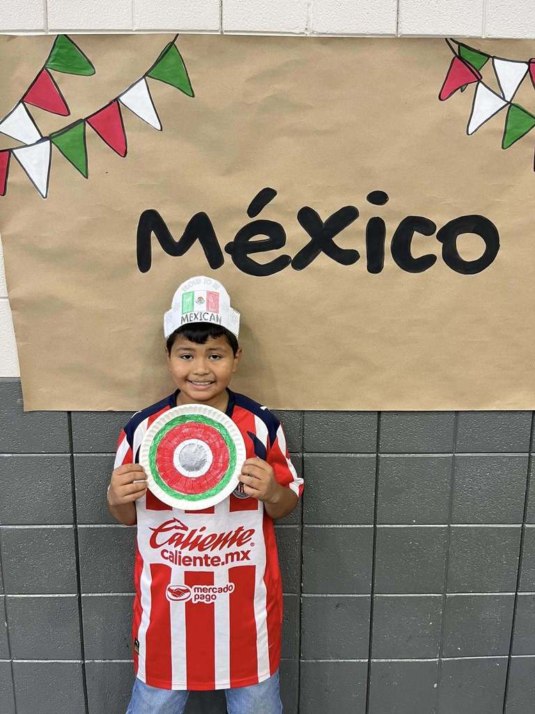 A student stands in front of a sign that says "Mexico" and holds a hand-made Mexican mirror.
