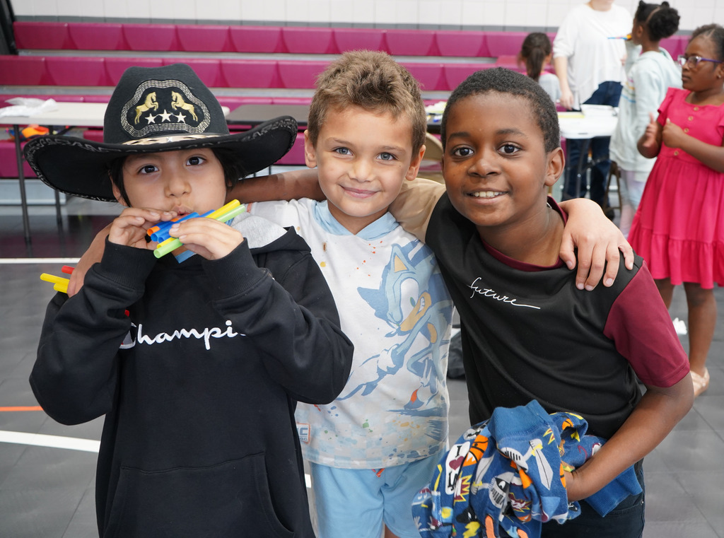 Students stand together, smiling for a photo in a school gym. One student is wearing a cowboy hat and playing a hand-made pan flute.