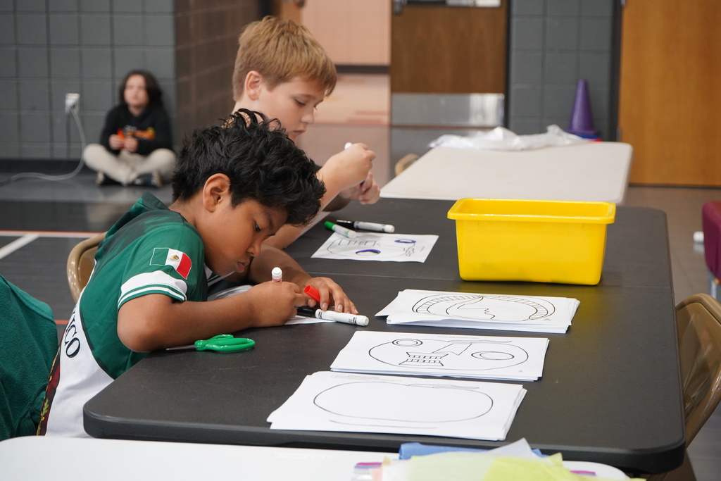Students sit at a table and color with markers.