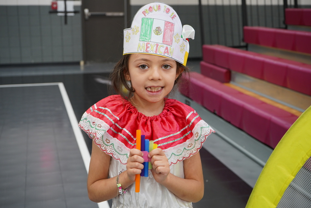 A student stands in a school gym and smiles while holding a hand-made pan flute.
