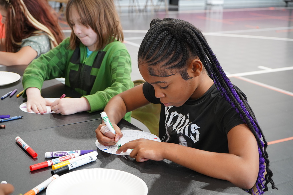 Students sit at a table and color with markers.