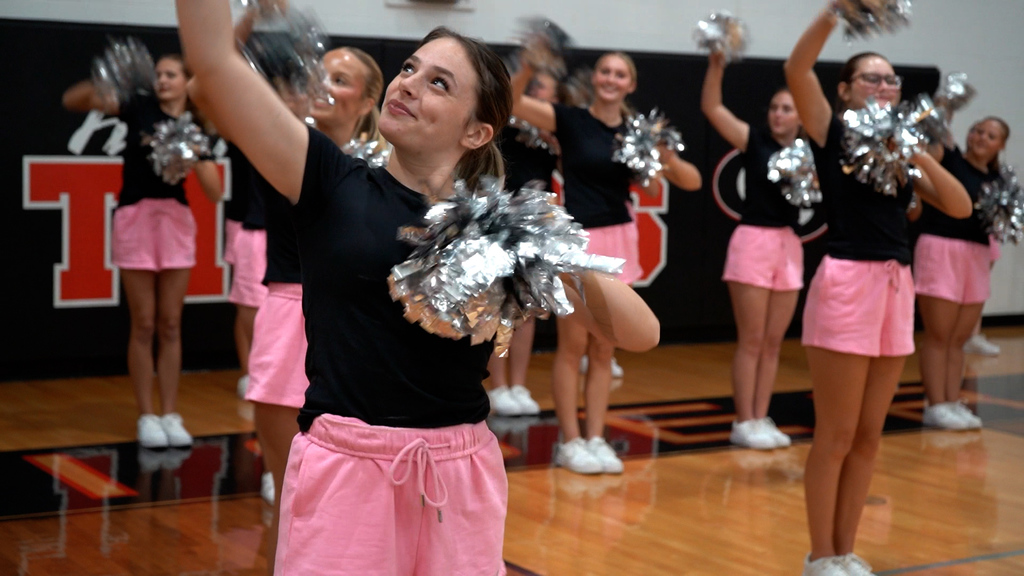 Girls wearing black shirts and pink shorts hold pom poms and smile while performing.