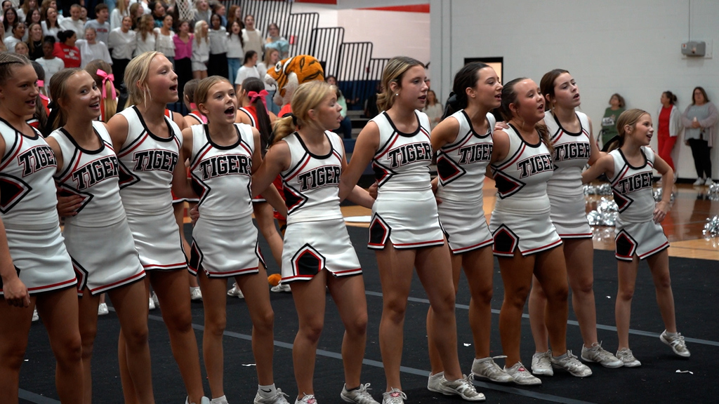Cheerleaders stand together and sing in a school gym.