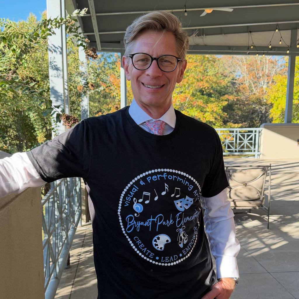 Alabama State Superintendent Dr. Eric Mackey stands on a porch outside. He wears a T-Shirt over a button down and tie. The T-Shirt says " Visual & Performing Arts Bryant Park Elementary Create Lead Empower."