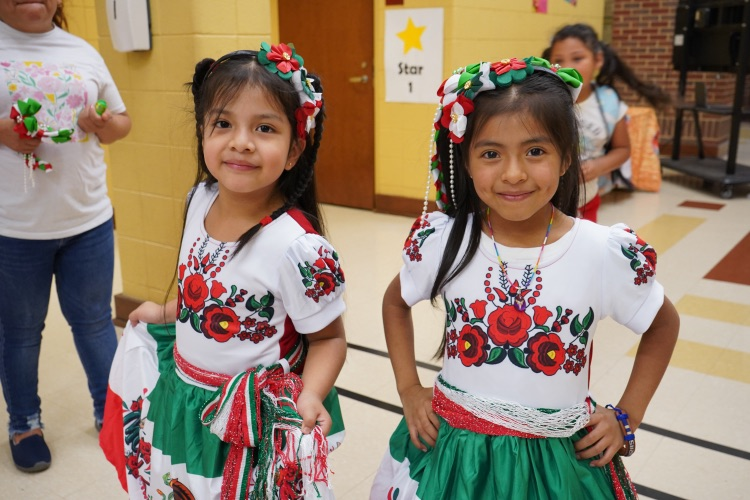 Two little girls wearing matching dresses smile for a picture in a school hallway.