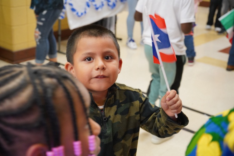 A little boy waves a flag while standing in a school hallway.