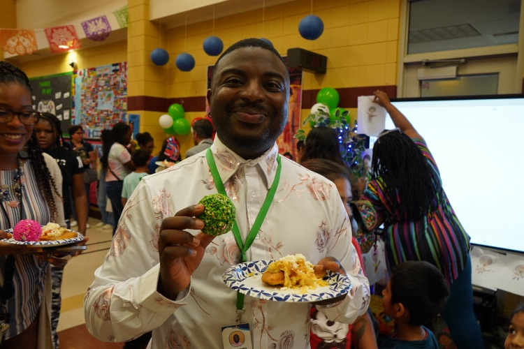 A man smiles while standing in a school hallway, holding a plate of food.