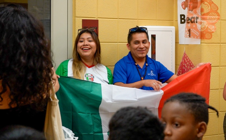 Two people stand and smile in a school hallway while holding up a flag.