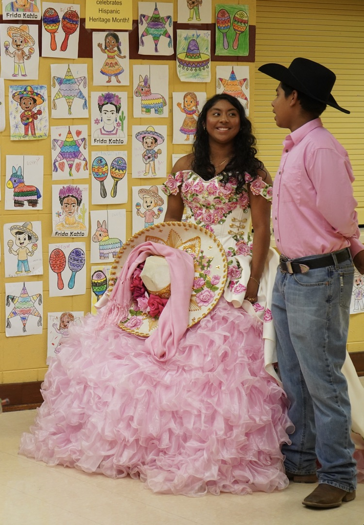 A teen girl and teen boy stand in a school hallway. The girl is wearing a pink dress and the boy is wearing a pink button up shirt, jeans and a hat.