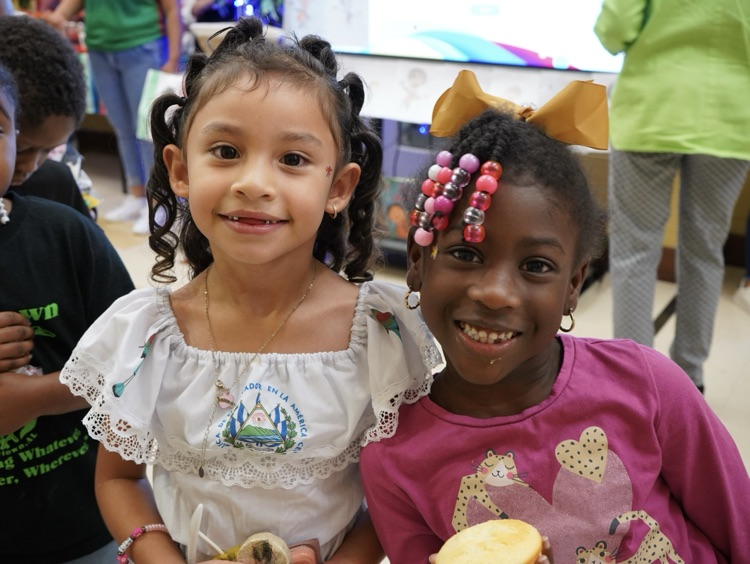 Two little girls stand in a school hallway and smile. They both hold treats in their hands.