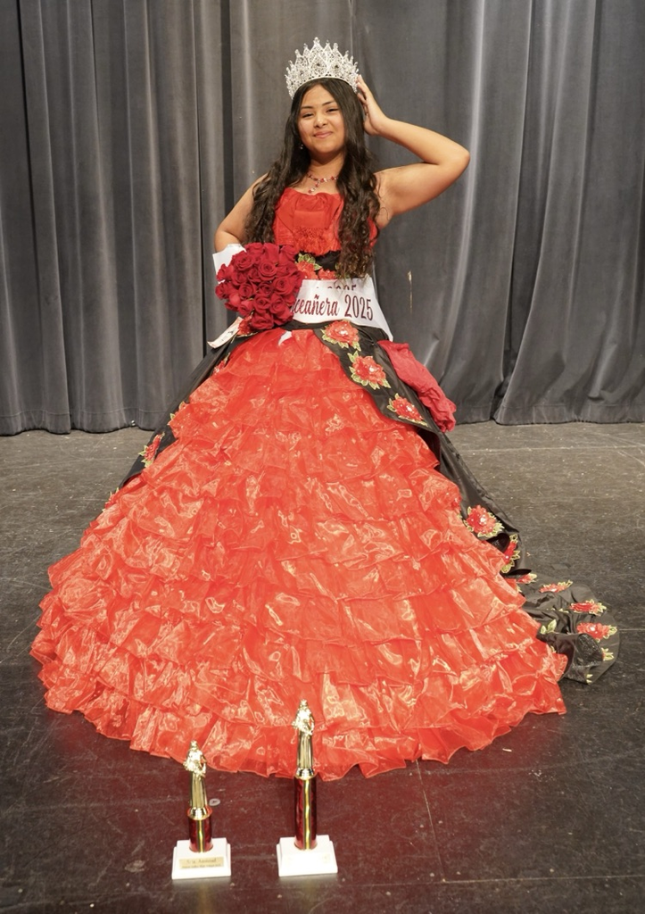 A student wearing a Quinceañera dress with a crown and sashes stands on a stage and holds flowers.