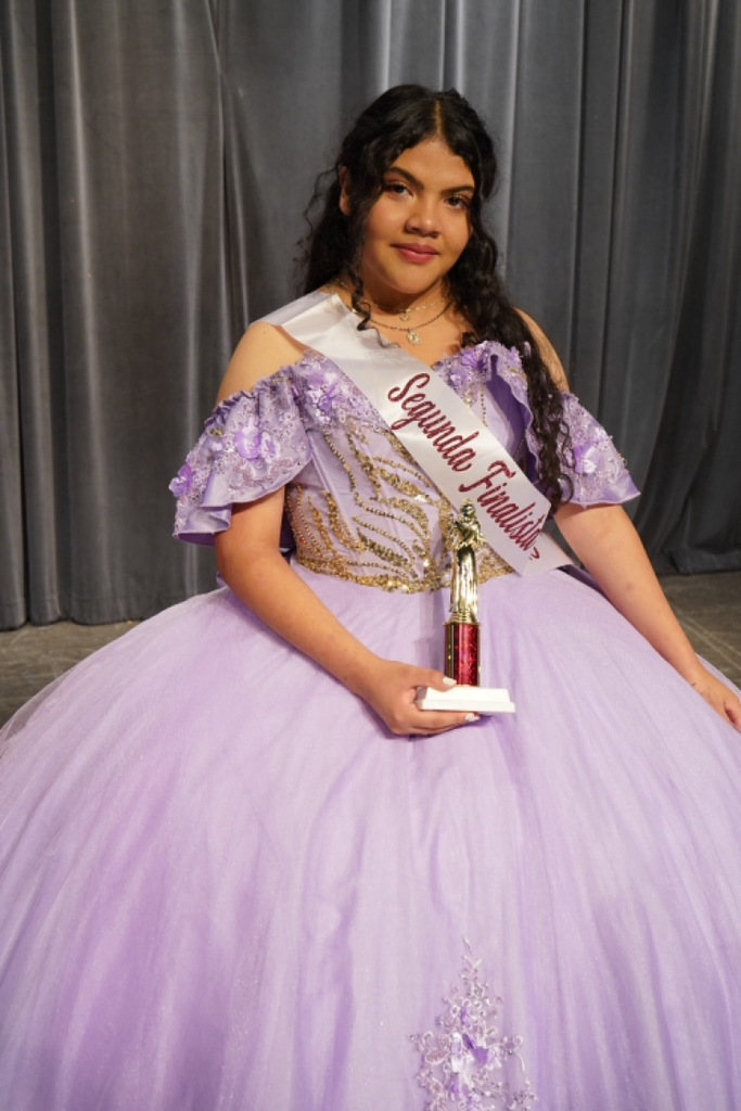A girl wearing a purple Quinceañera dress holds a trophy and smiles.