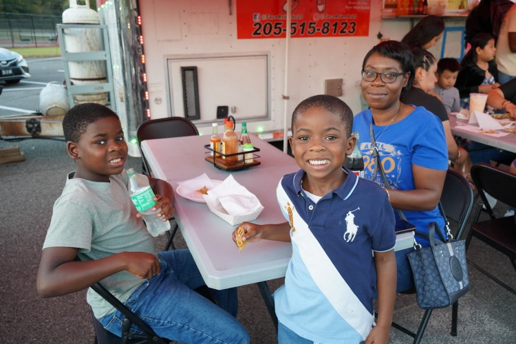 A family sits and stands together and smiles while eating at a food truck at a Hispanic Heritage Festival.