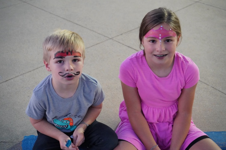 Two children with face paint smile and sit on a sidewalk.