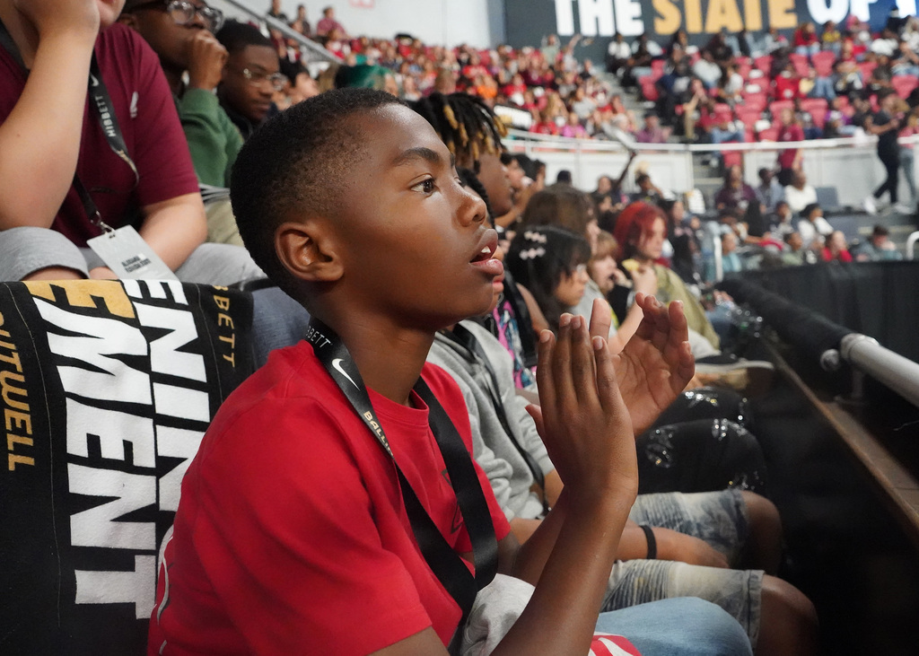 This is a close up photo of a male student in a red shirt clapping while watching a basketball game.