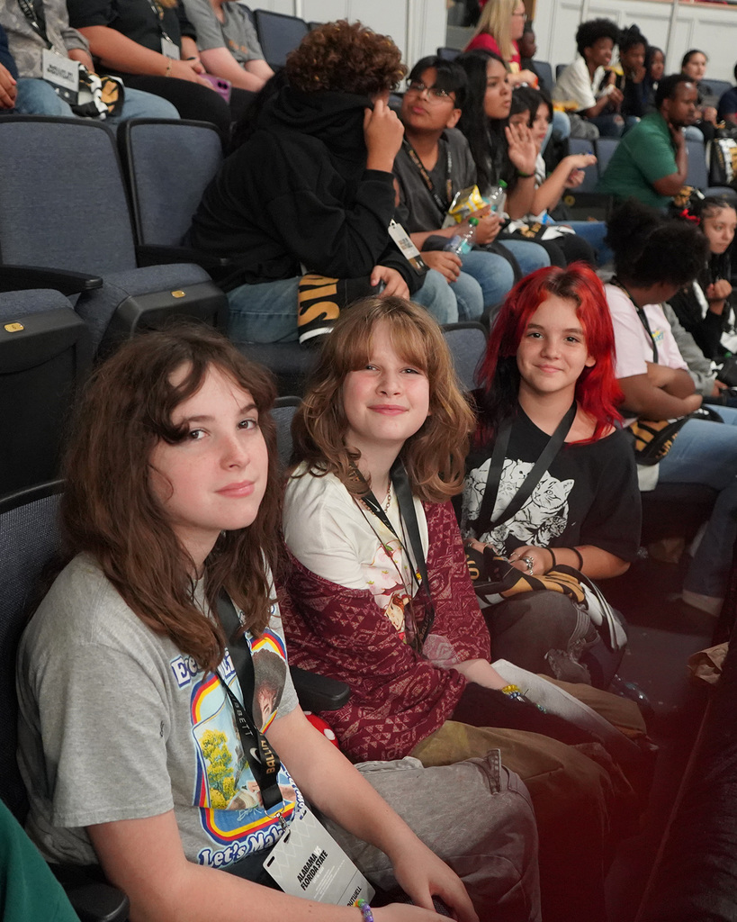 Three female students pose for a picture during a basketball game. They are sitting in the crowd.