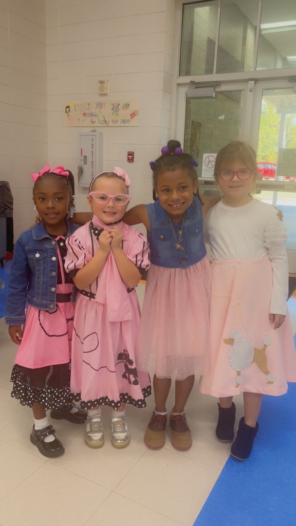 Four little girls wear 50s inspired outfits and pose together for a photo while standing inside a school hallway.