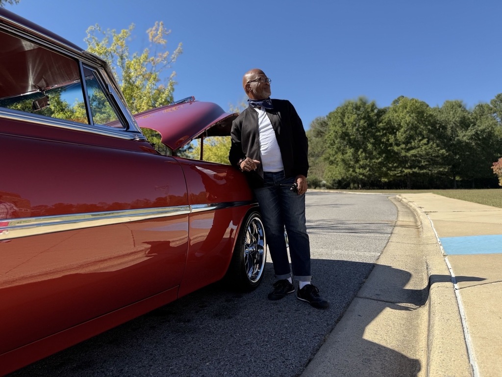 A man wearing a 50s inspired outfit stands next to a vintage car.