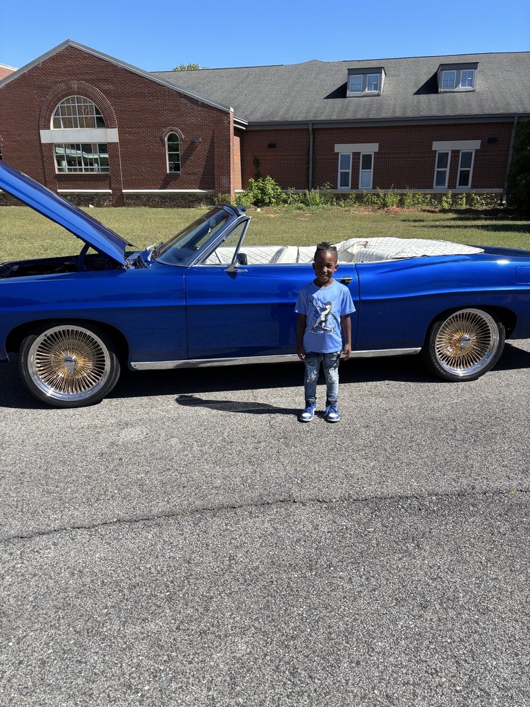 A little boy stands in front of a vintage car and smiles.