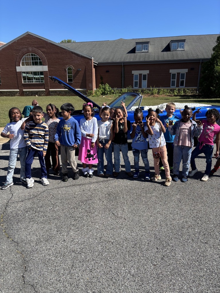 A group of students pose for a photo outside a school while standing in front of a vintage car.
