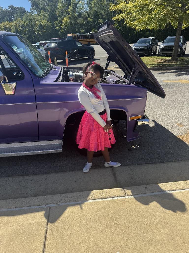 A girl wearing a 50s inspired outfit stands in front of a vintage car.