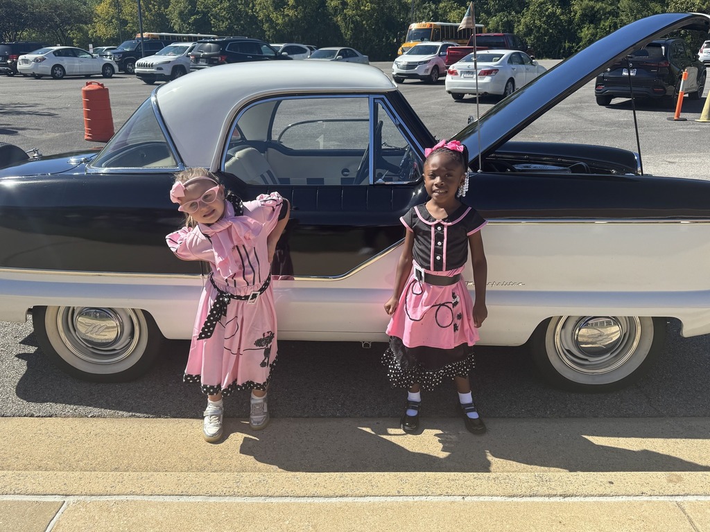 Two girls wearing 50s inspired outfits stand in front of a vintage car and smile.