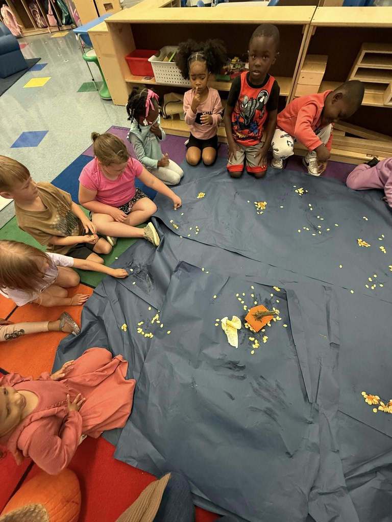 Students sit on the floor of a classroom. In front of them are several large pieces of black paper with pumpkin seeds sitting on them.