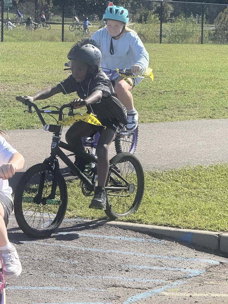 Students ride bicycles on grass and on a parking lot.