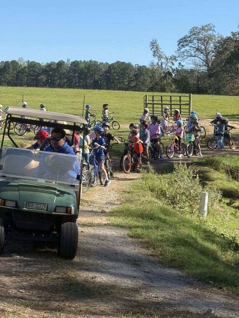 Students ride bikes through a grassy area. In front of the students is a man driving a golf cart.