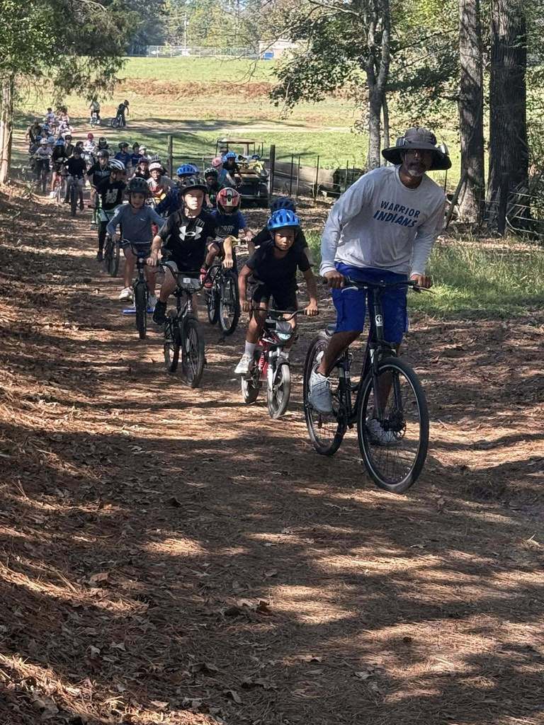 Students and an adult ride bicycles through a wooded area.