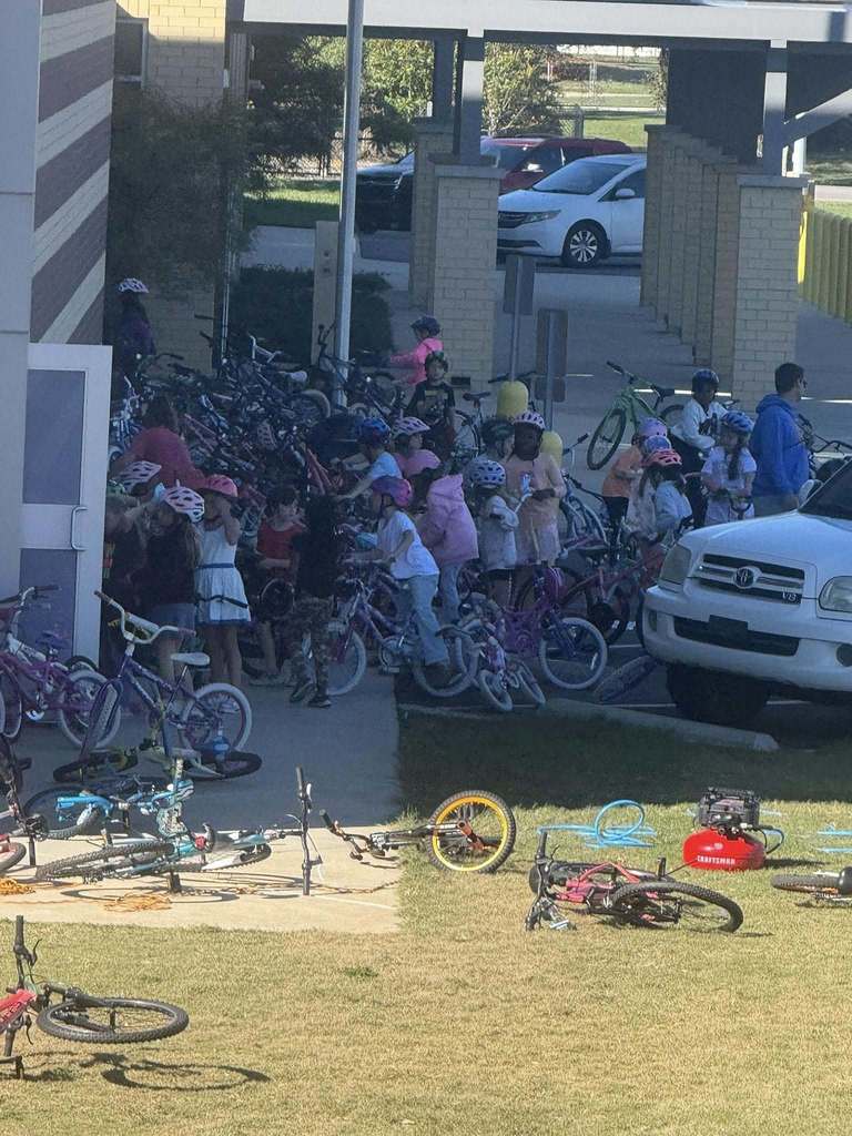 Many students stand on and next to bicylces outside a school.