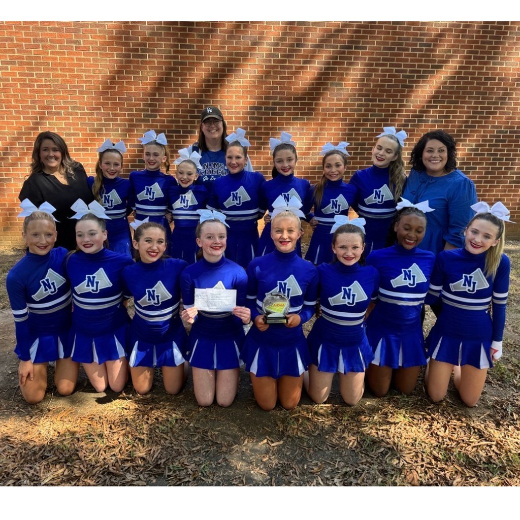North Jefferson Middle School cheerleaders and three adults pose togehter for a picture. Cheerleaders in the middle hold a piece of paper and a trophy.