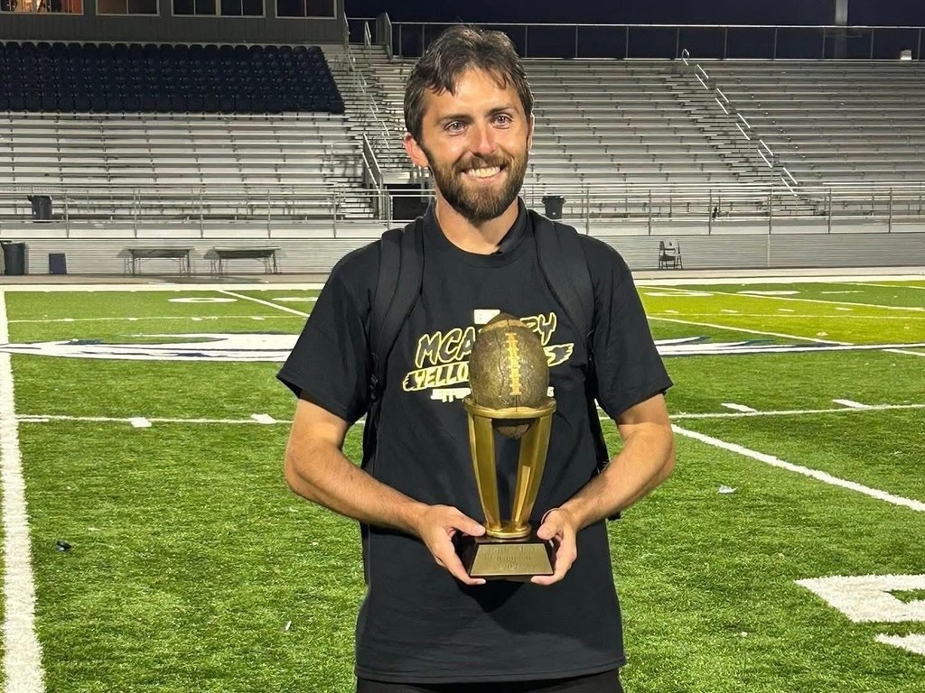 McAdory Middle School's football coach holds a trophy and smiles while standing on a football field at night.