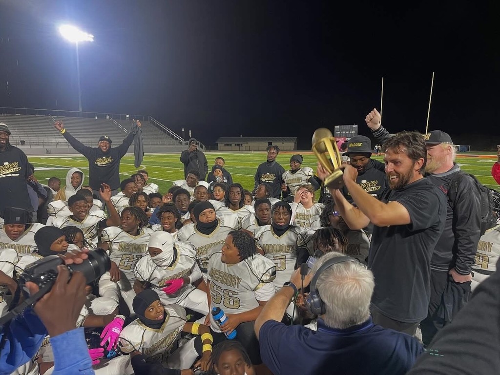 McAdory Middle School's football team and coaches sit and stand on a football field at night. The players and coaches look happy as the head coach holds a trophy up.