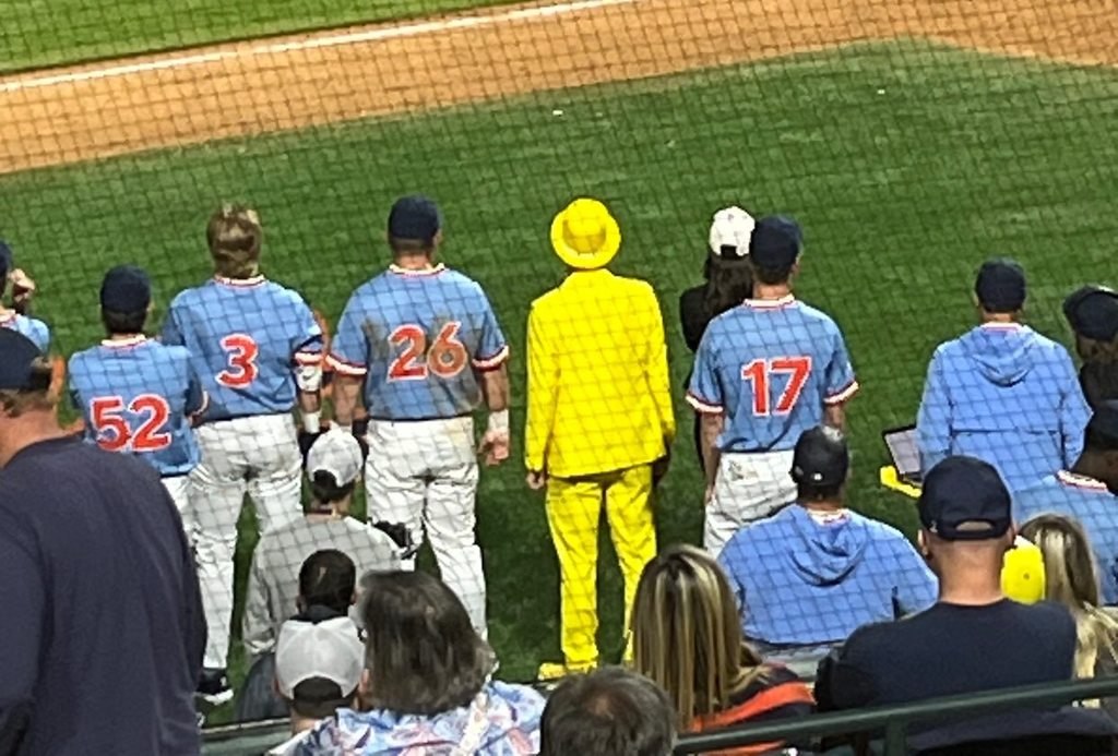 Baseball players and a man in a yellow suit stand on a baseball field.